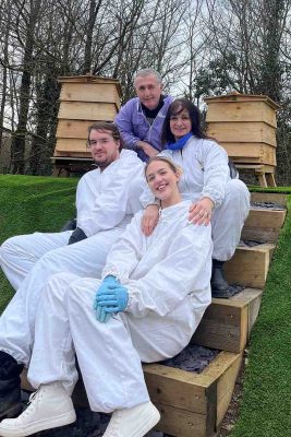 Four people in beekeeping suits posing happily on wooden steps in front of beehives, showcasing their readiness for beekeeping activities