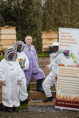 Three people in beekeeping suits standing next to a banner promoting corporate well-being and environmental renewal through beekeeping.