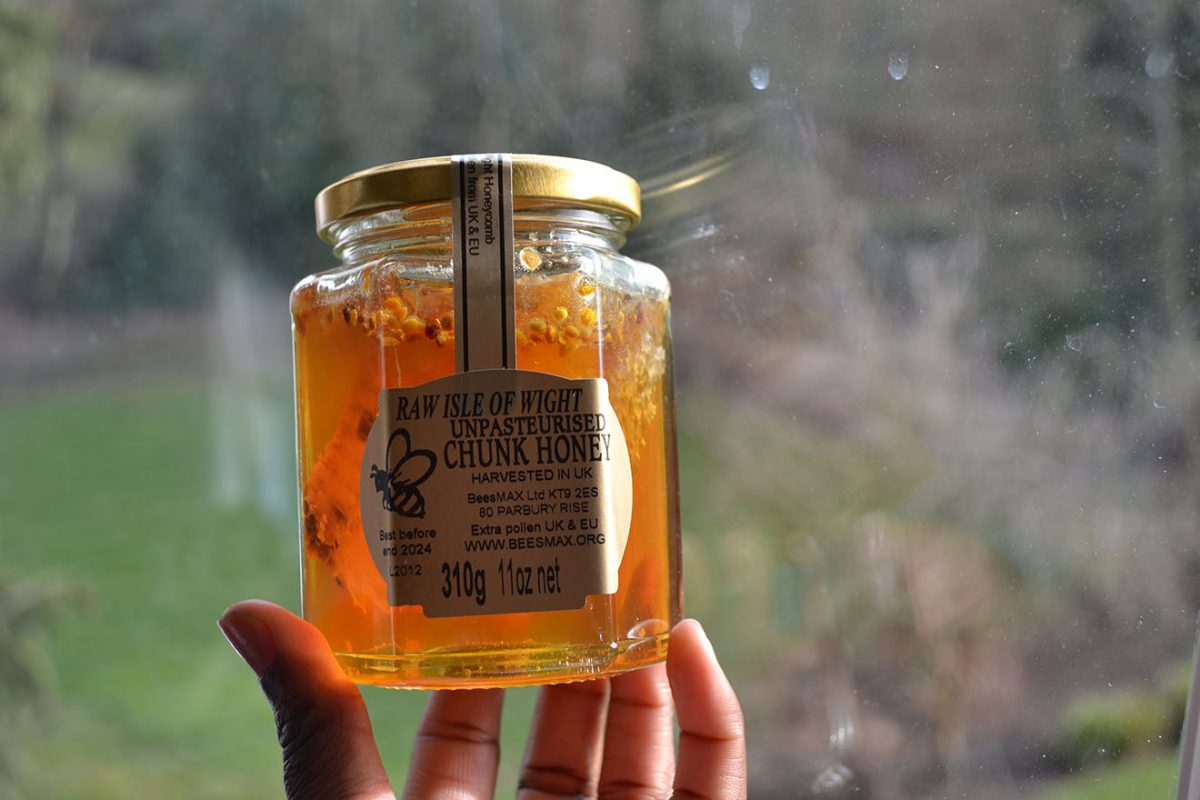 Someone's hand holding up a jar of raw 'BeesMAX' chunk honey against a window with the daylight clearly showing honeycomb inside the jar in the joney.